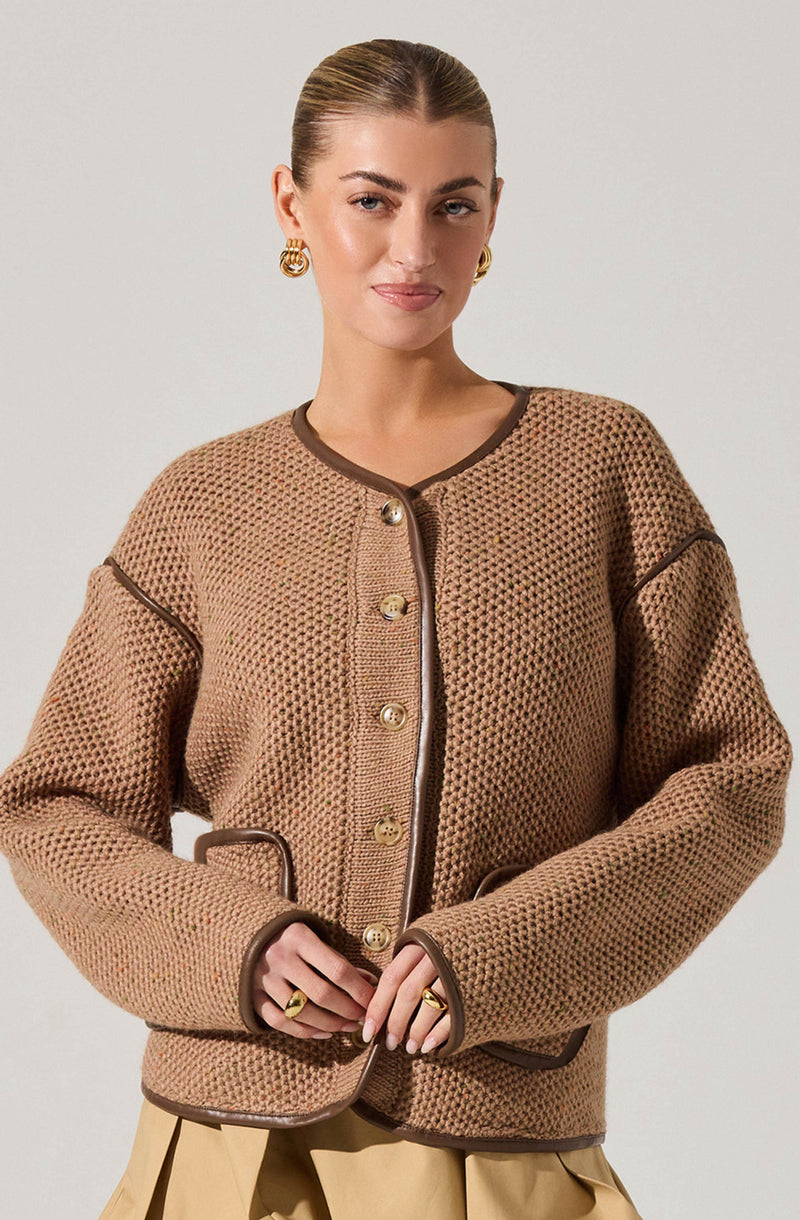 A woman is wearing a beige, textured knit jacket with dark brown trim along the collar, front opening, pockets, and sleeve seams. The jacket features gold buttons down the front and pockets, with the trim adding contrast.