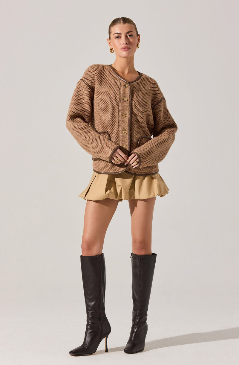 A woman is wearing an oversized, textured, brown knitted cardigan with large buttons down the front. She is also wearing a beige ruffled mini skirt and tall, black leather high-heeled boots.