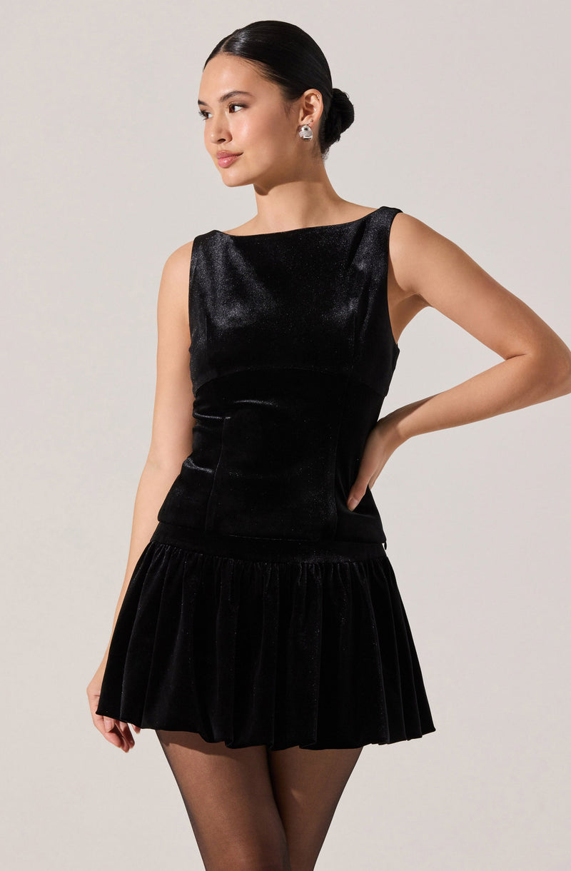 A woman is wearing a sleeveless black velvet dress with a fitted bodice and a rounded neckline. The dress has a short, pleated skirt that adds volume and texture to the outfit. The woman is accessorized with large, round earrings and has her hair styled in a sleek, low bun.