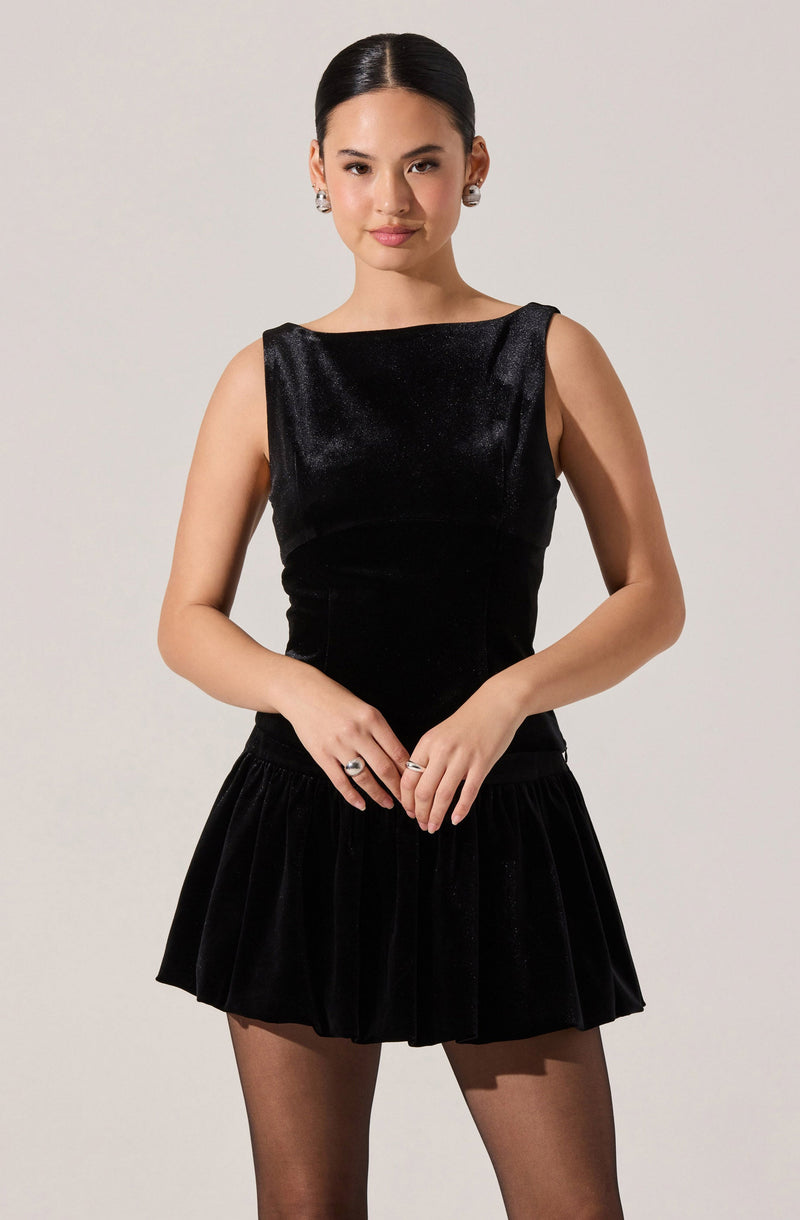 A woman is wearing a sleeveless black top made of a shiny, textured fabric with a round neckline. She pairs it with a black velvet skirt that features a ruffled hem and a drawstring waist. She accessorizes with large earrings and rings on her fingers.