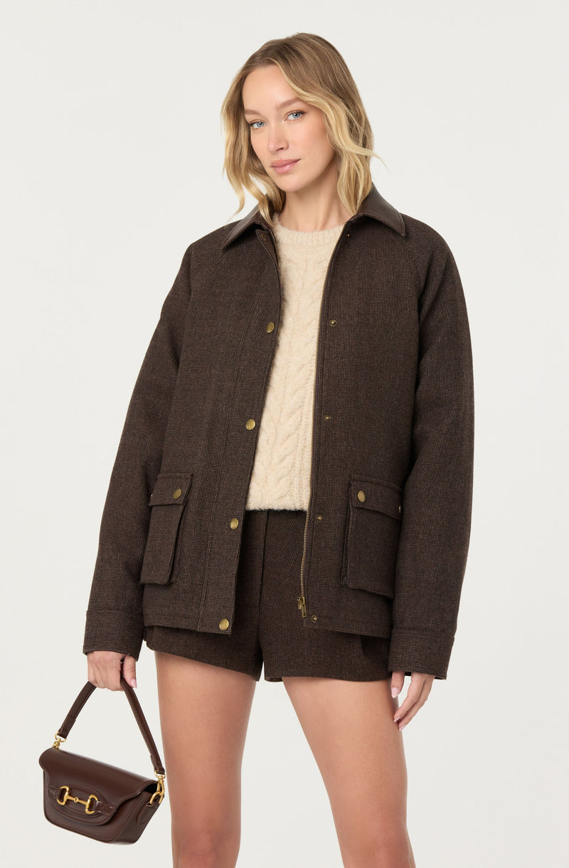 A woman is wearing a dark brown, textured jacket with a collar and gold buttons. Underneath, she has on a cream-colored, cable-knit sweater. She is also wearing matching dark brown shorts and is holding a small brown handbag with gold hardware.