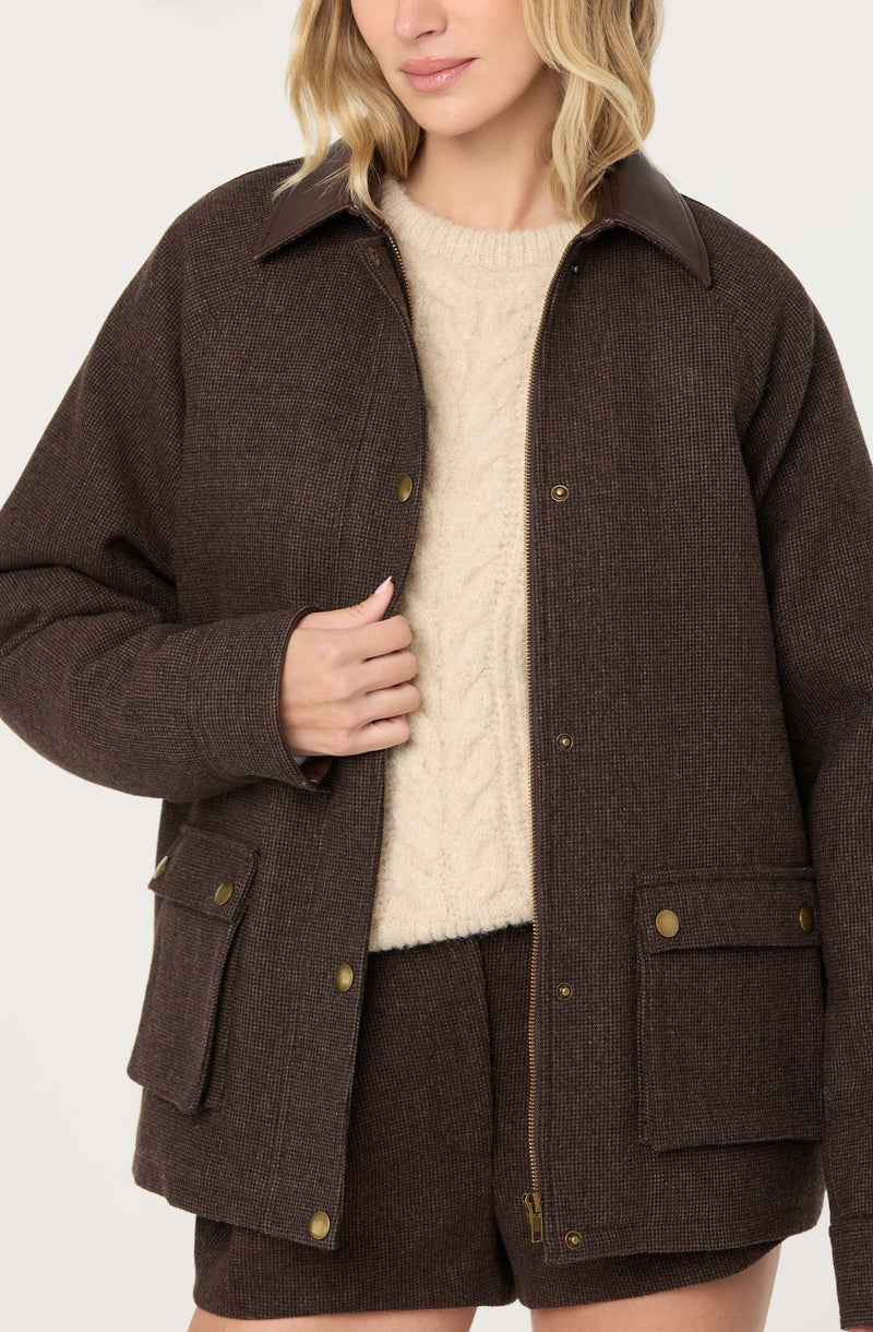 A woman is wearing a dark brown, textured jacket with a collar made of a different material, possibly leather. Underneath, she has on a cream-colored, cable-knit sweater. The jacket features gold buttons and has large patch pockets with button flaps on the front. She is also wearing matching dark brown textured shorts.