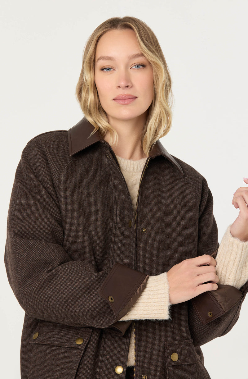 A woman is wearing a dark brown, textured, zip-up jacket with a pointed collar and leather-like accents on the cuffs and pockets. Underneath, she has on a light beige, ribbed knit sweater with long sleeves.