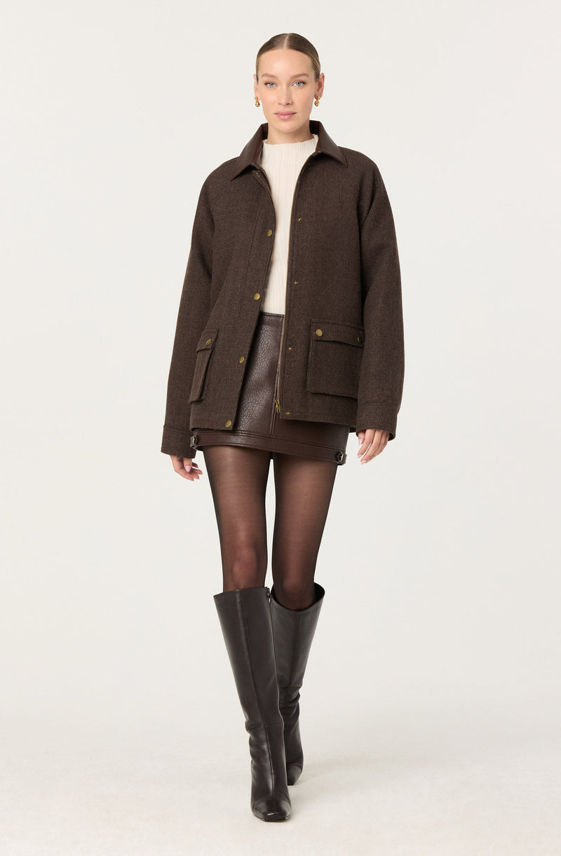 A woman is wearing a dark brown, oversized, collared jacket with gold buttons and large front pockets. Underneath, she has on a cream-colored knitted sweater. She is also wearing a high-waisted, dark brown leather mini skirt with a front zipper and snap button details. Her outfit is completed with sheer black tights and tall black leather boots that reach just below her knees.