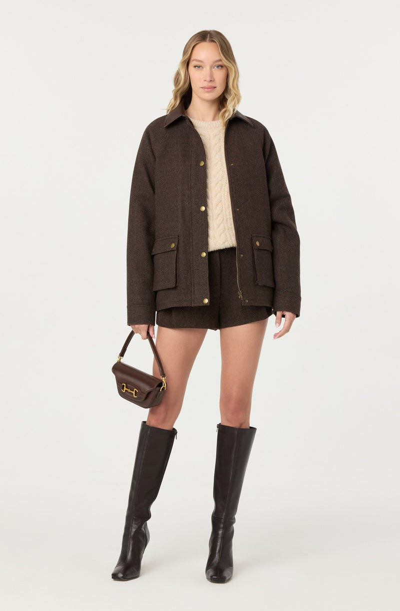 A woman is wearing a dark brown woolen jacket with gold buttons, a cream cable knit sweater underneath, and dark brown shorts. She is carrying a small dark brown handbag with a gold clasp and is wearing tall black leather boots that reach just below the knee.