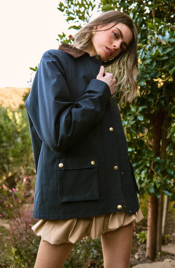 A woman is wearing a navy blue jacket with large gold buttons and a brown collar. The jacket has two front pockets with gold button flaps. She is also wearing beige, puffy shorts with a gathered, bubble-like appearance. The background shows lush green foliage and a partly cloudy sky.