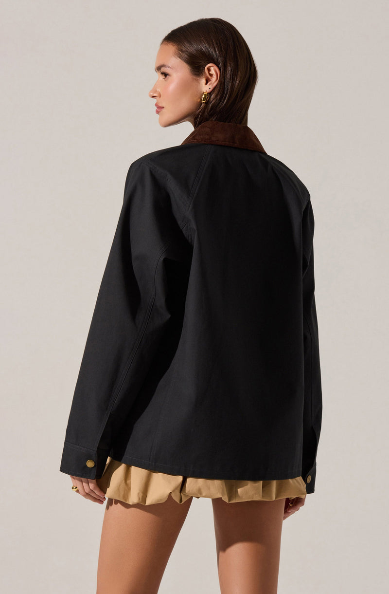 A woman wearing a black jacket with a brown collar, featuring long sleeves with buttoned cuffs, and beige shorts with a ruffled hem.