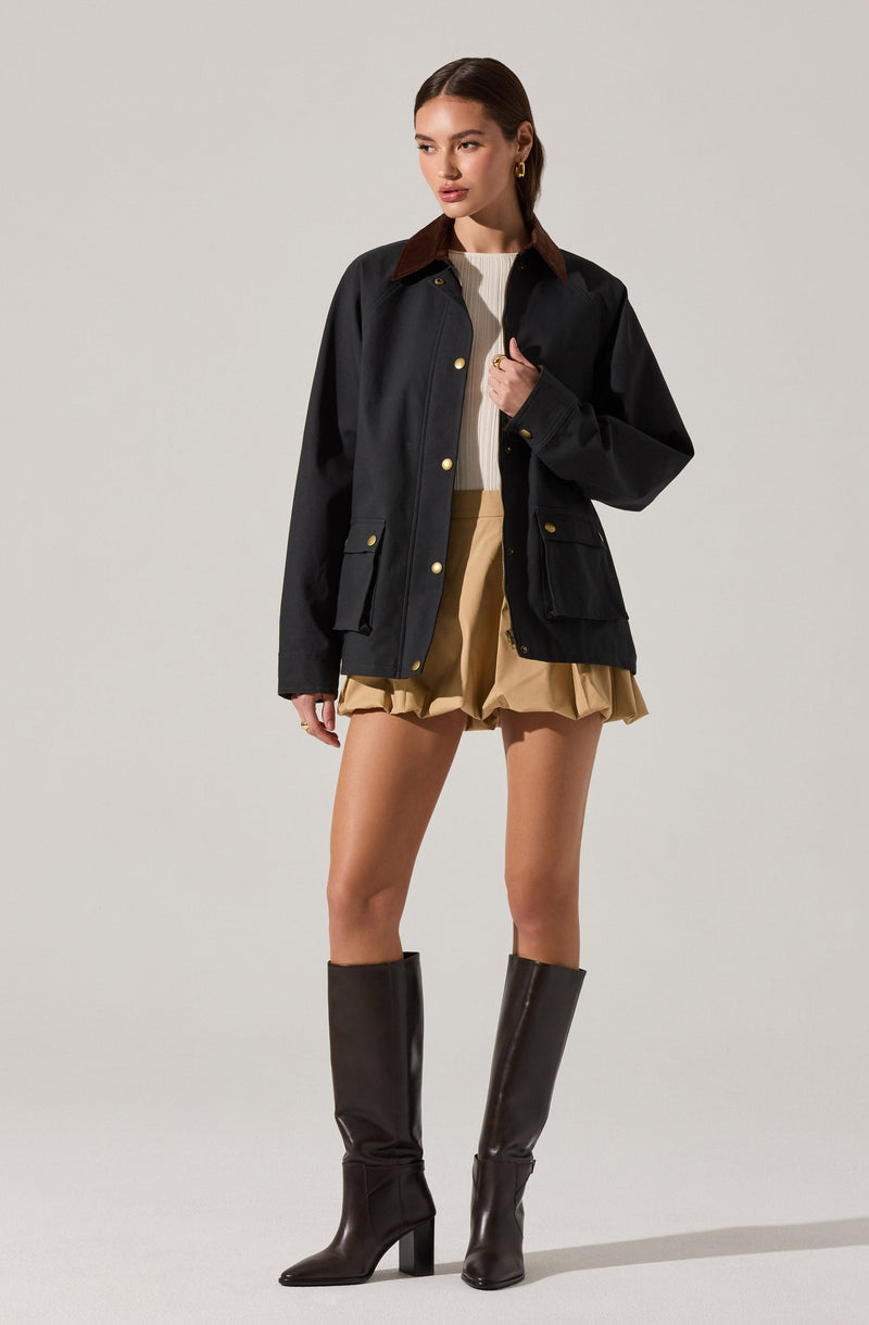 A woman is wearing a black jacket with gold buttons, a brown collar, and multiple large pockets. Underneath, she has on a cream-colored, ribbed, crew-neck sweater. She is also wearing beige shorts with gathered ruffled hems. She has on knee-high black leather boots with block heels.