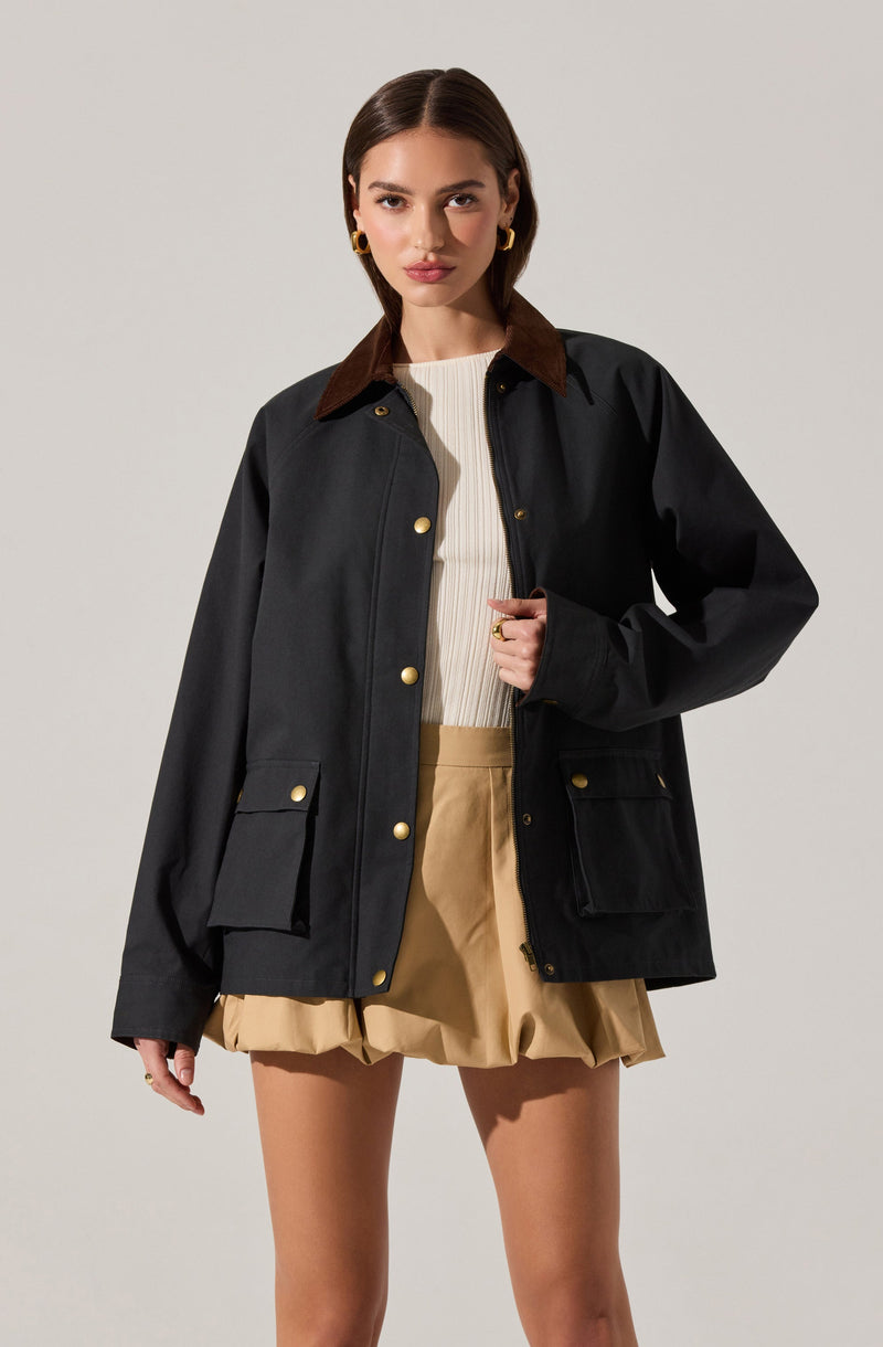 A woman is wearing a black jacket with a brown collar, featuring gold buttons and large front pockets. Underneath, she has on a white ribbed top. She is also wearing beige shorts with a gathered, bubble-like hem.
