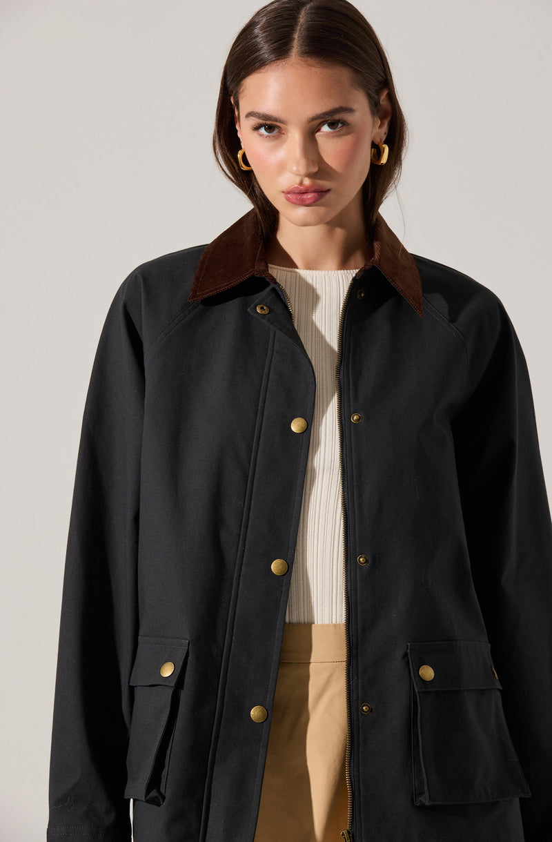 A woman is wearing a black jacket with a brown corduroy collar, gold snap buttons, and front flap pockets. Underneath, she has on a cream ribbed sweater and beige pants. She has gold hoop earrings and has styled her hair in a sleek, straight manner.