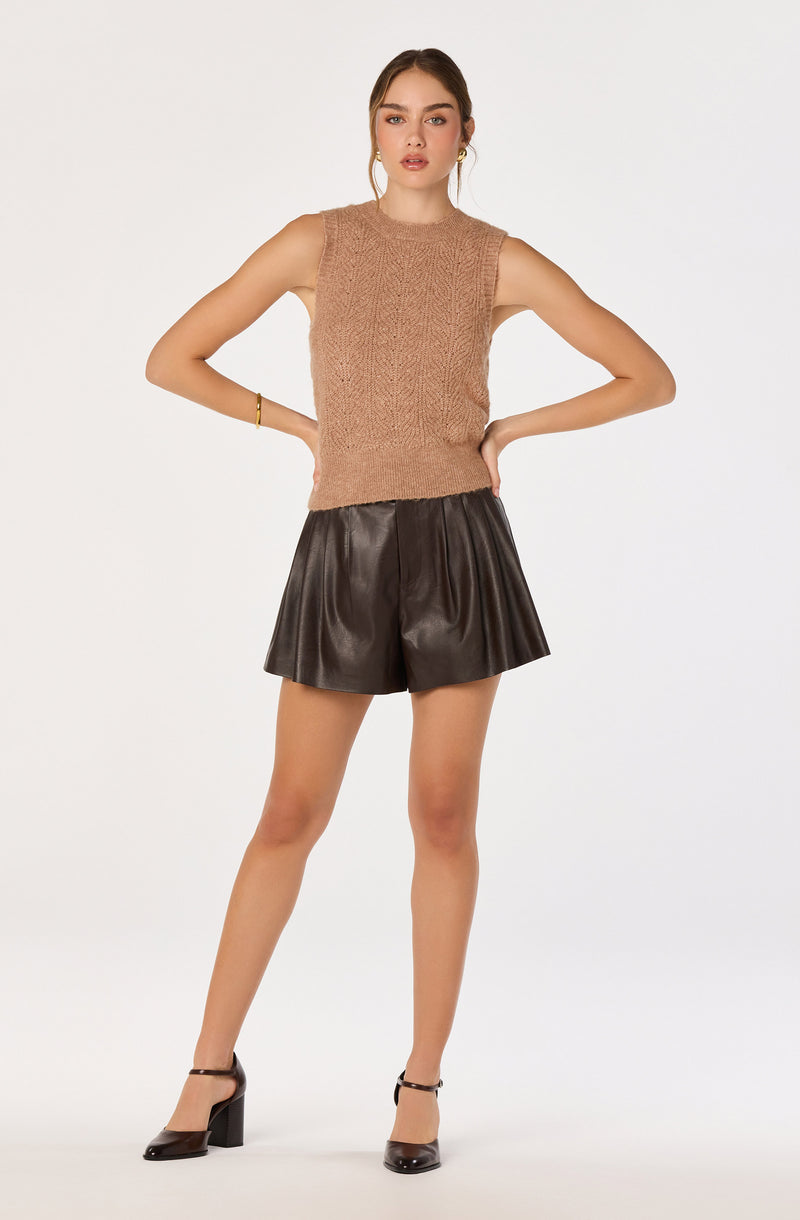 Woman in sleeveless beige knit top, black leather shorts, and black high heels.