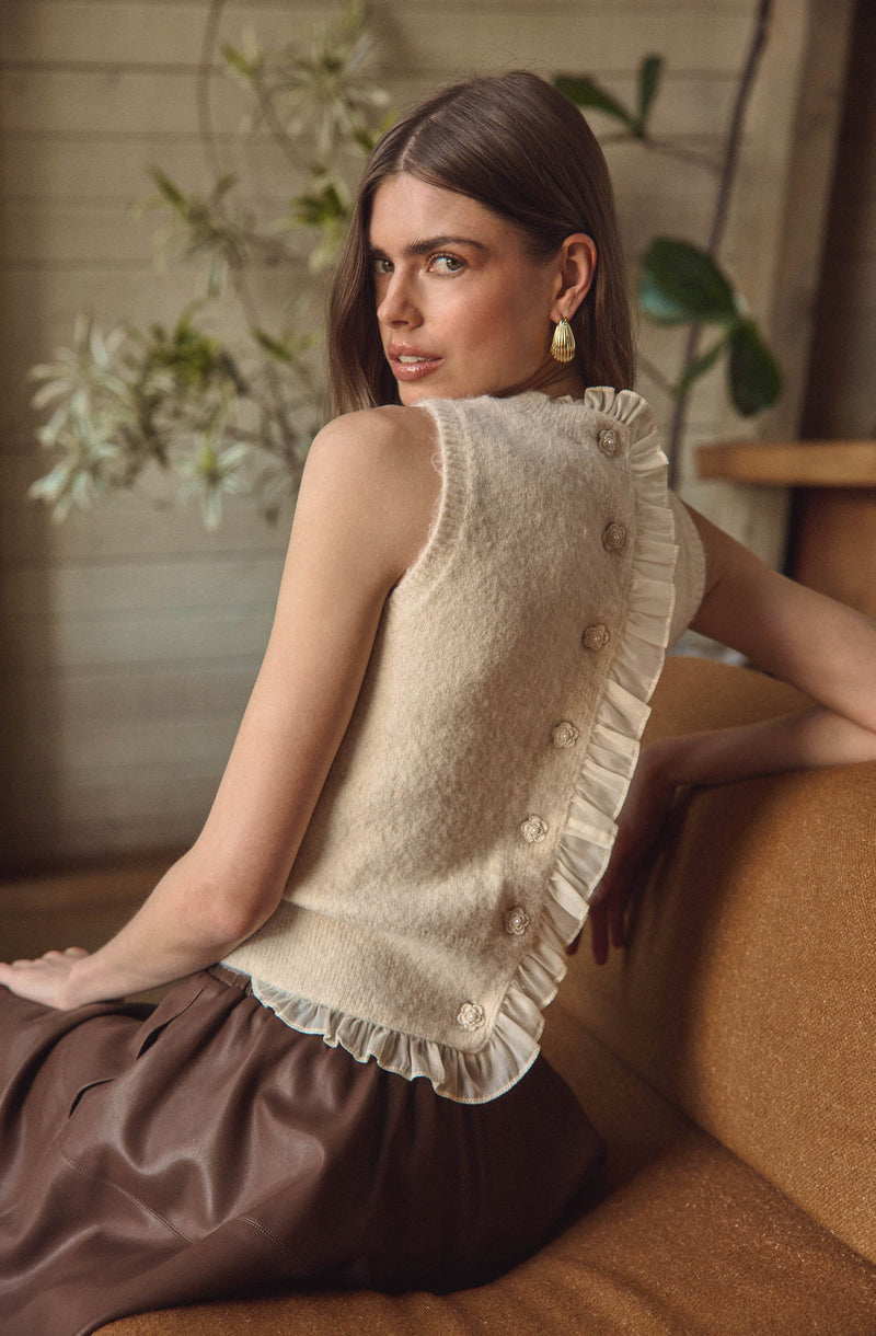 A woman is wearing a sleeveless, cream-colored top made of a fuzzy knit fabric. The top features a row of decorative buttons along one side, with ruffled trim alongside the buttons. The top appears to have a slightly ruffled hem.