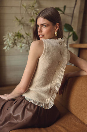 A woman is wearing a sleeveless, cream-colored top made of a fuzzy knit fabric. The top features a row of decorative buttons along one side, with ruffled trim alongside the buttons. The top appears to have a slightly ruffled hem.