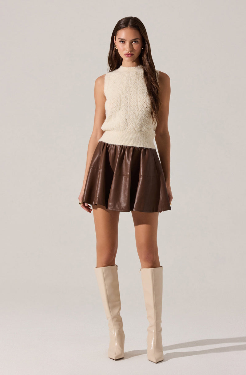 A woman is wearing a sleeveless, high-neck, cream-colored knit top and a short, voluminous brown leather skirt. She has on tall, cream-colored boots that reach just below her knees.
