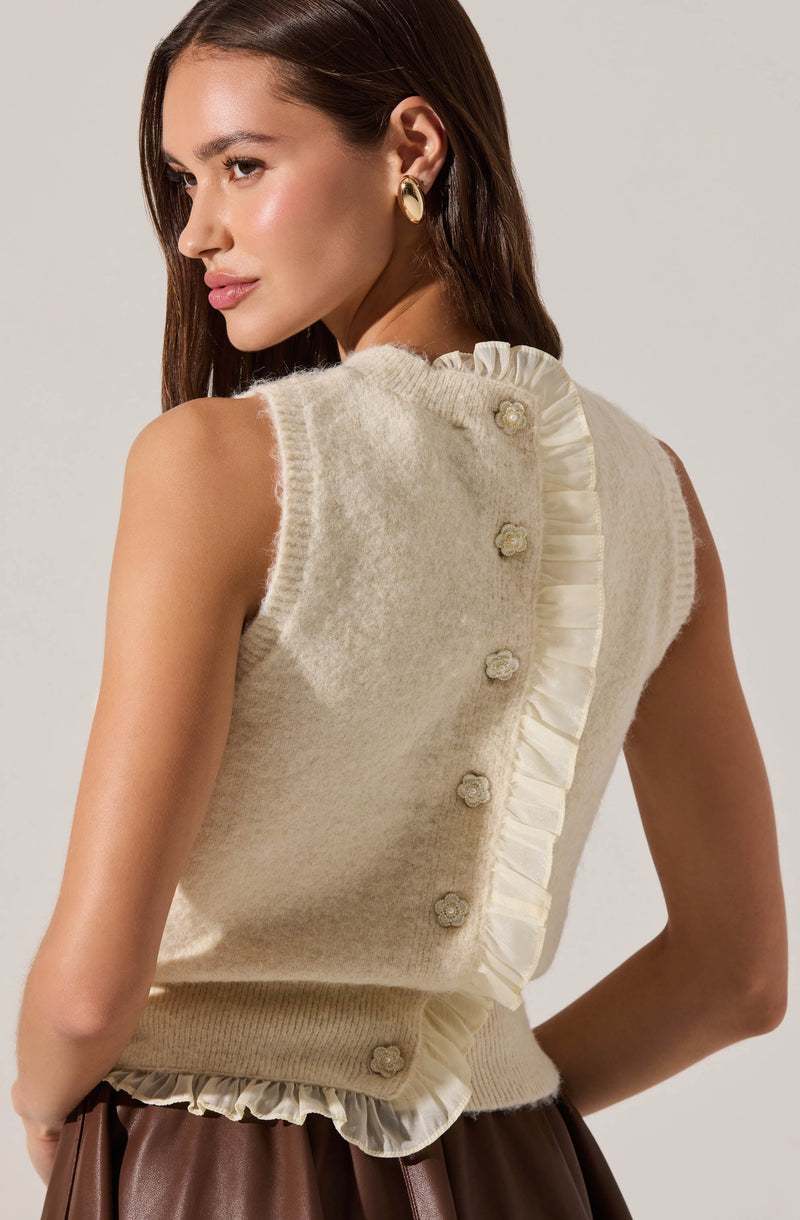 A woman is wearing a sleeveless, cream-colored knitted vest with a soft, fuzzy texture. The vest features decorative fabric ruffles along the back and neckline, with small, fabric flower buttons running down the back. She pairs it with a brown skirt or pants and accessorizes with large, gold hoop earrings.