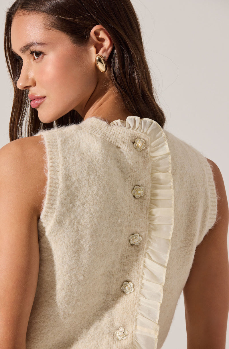 A woman with long brown hair wears a sleeveless, cream-colored sweater with a textured, fuzzy fabric. The sweater features decorative buttons shaped like flowers and has a ruffled satin trim running vertically along the back. She accessorizes with gold oval earrings.