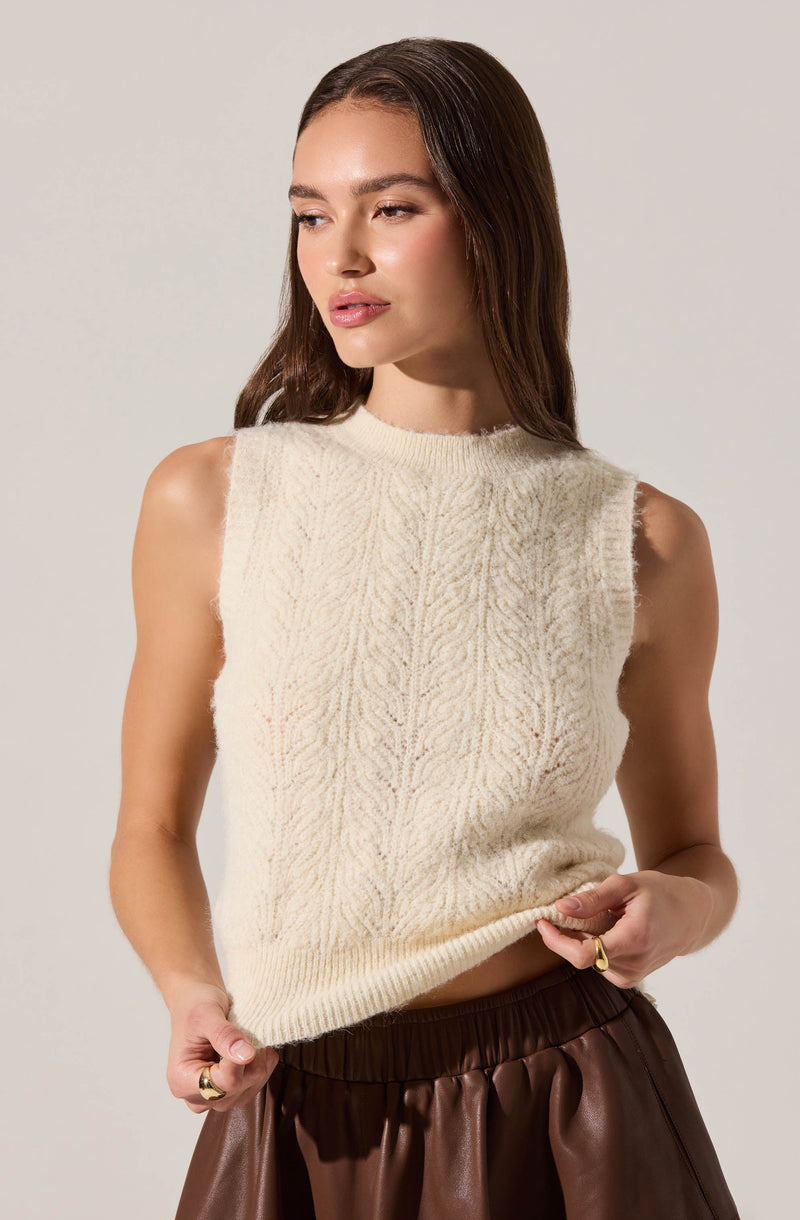 A woman is wearing a sleeveless, cream-colored knitted vest with a subtle textured pattern. The vest has a ribbed neckline and hem, and the armholes have a slightly frayed edge. She pairs it with dark brown, shiny, high-waisted pants.