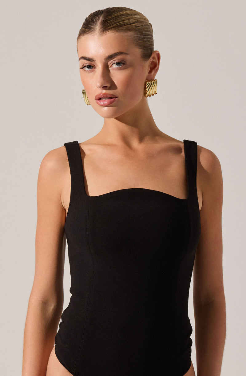 A woman is wearing a black sleeveless top with wide straps that sit on her shoulders. The top has a square neckline with a slight curve across the bust. She is accessorized with large, gold, spiral-shaped earrings.