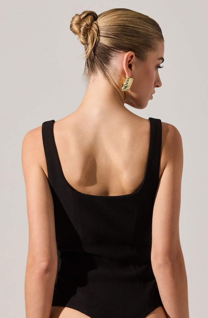 A woman is wearing a sleeveless, black bodysuit with wide straps and a square neckline. Her hair is styled in a tight bun, and she has large, gold, crescent-shaped earrings.