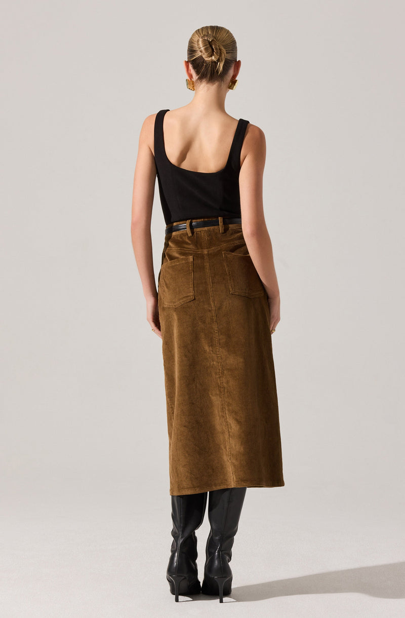A woman is wearing a black sleeveless top with wide straps, tucked into a high-waisted, long brown corduroy skirt that reaches her ankles. She accessorizes with large gold earrings and is also wearing black high-heeled boots.