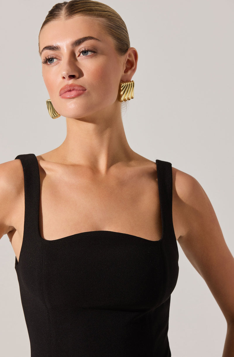 A woman is wearing a black sleeveless dress with wide straps and a fitted bodice. She also has large, gold, sculptural earrings with a ribbed, wavy design. Her hair is slicked back, and she has a neutral makeup look.