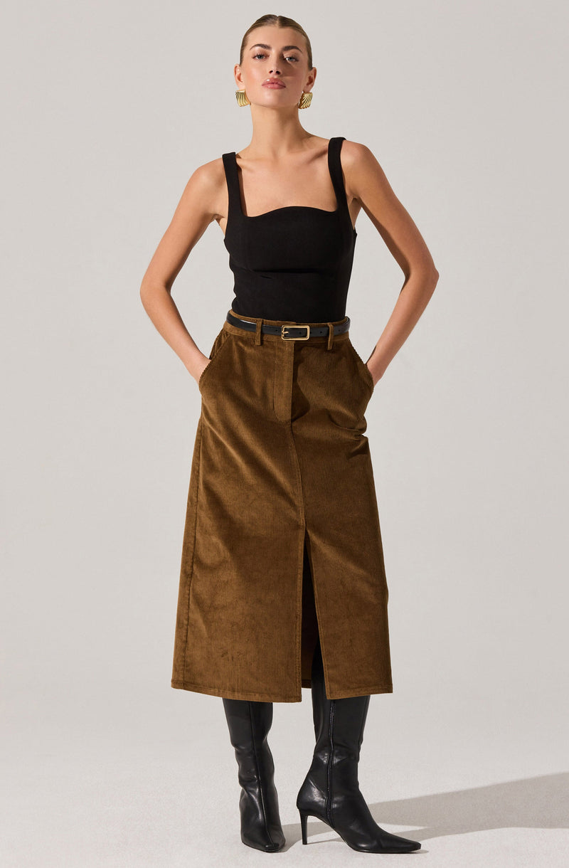 A woman is wearing a black sleeveless top with wide shoulder straps and a square neckline. She pairs it with a high-waisted, long brown skirt that has a front slit and is secured with a black belt. She completes her outfit with black knee-high boots and gold hoop earrings.