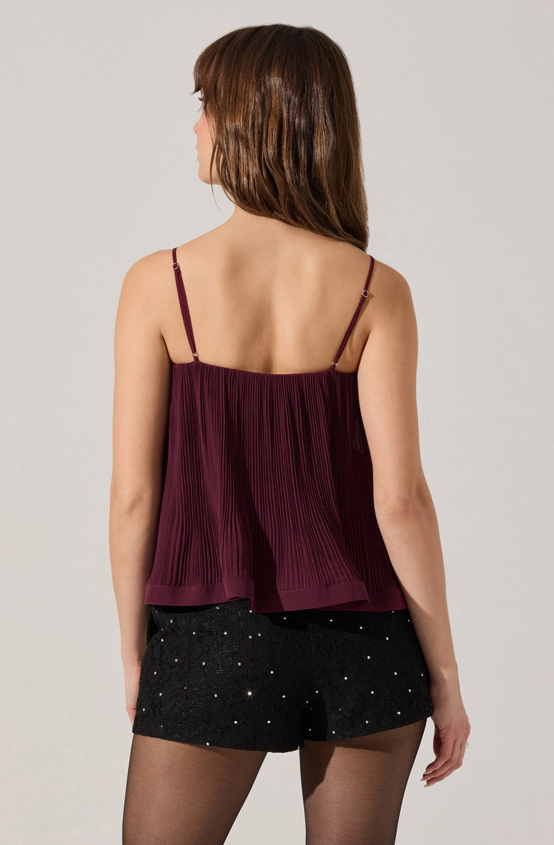 A woman is wearing a sleeveless, burgundy top with thin adjustable spaghetti straps. The top features folded pleats and a flowing, relaxed fit. She pairs it with black shorts that are decorated with small, scattered rhinestones or glitter embellishments.