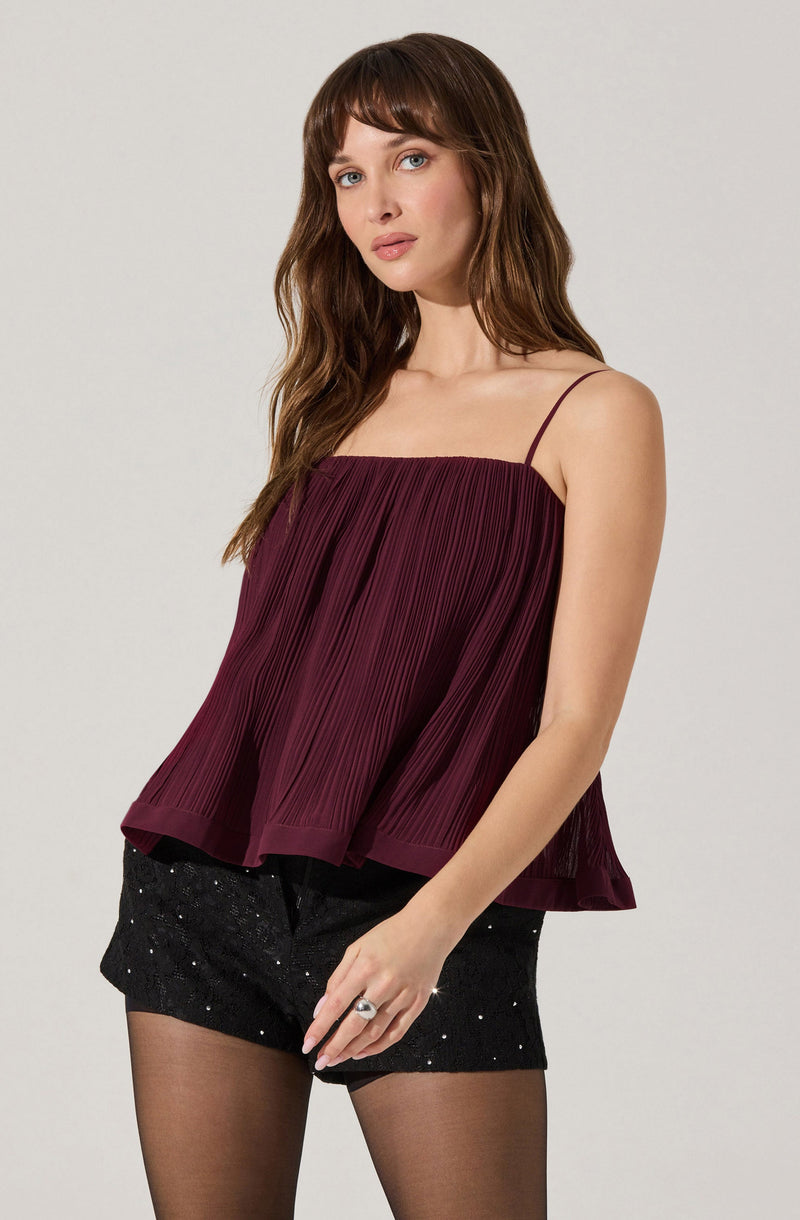 A woman is wearing a sleeveless, burgundy top with thin spaghetti straps. The top features a pleated, flowy design and a slightly flared hem. She is also wearing black shorts with a high waist, decorated with small, shiny embellishments, and sheer black stockings.