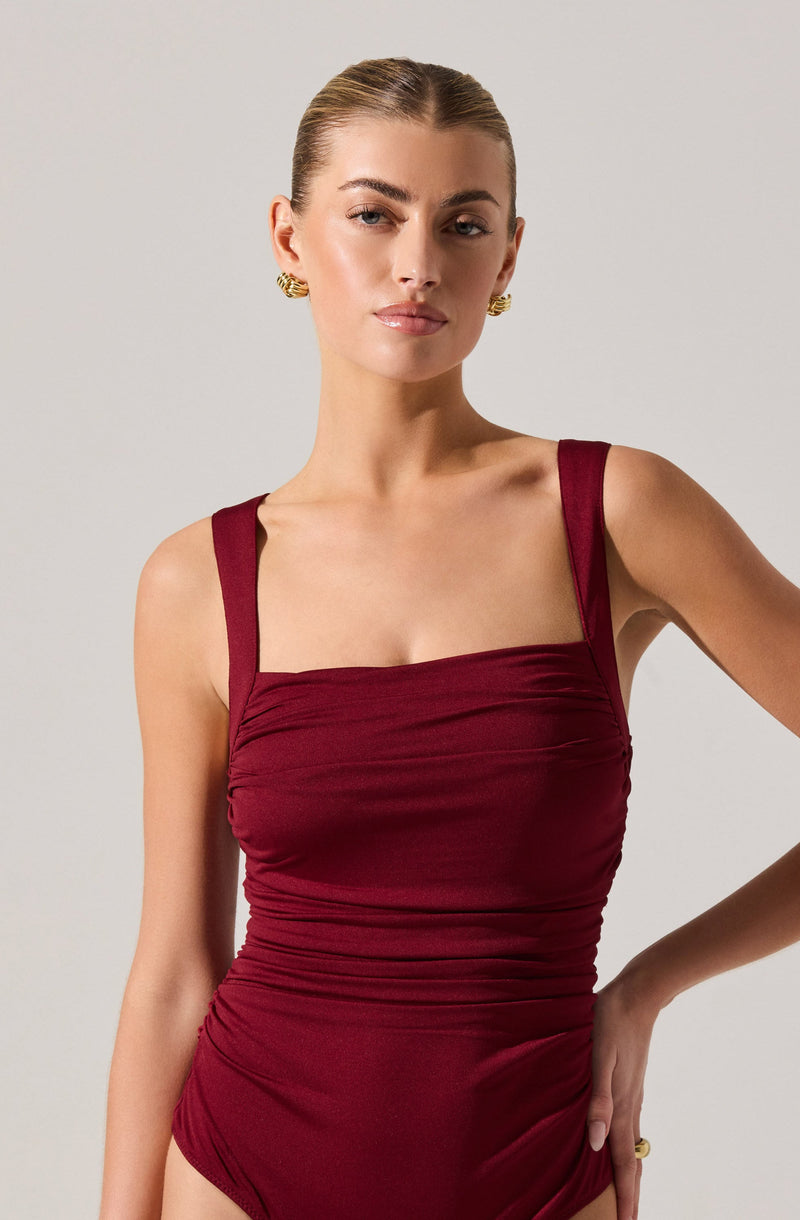 A woman is wearing a sleeveless, form-fitting maroon dress with thick straps and a ruched bodice. She has her hair pulled back and is accessorized with gold earrings and a gold ring on her right hand.