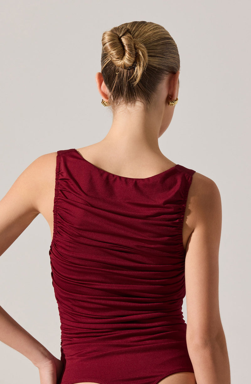 A woman is wearing a sleeveless, deep red or burgundy dress with a rounded neckline. The dress features a gathered, ruched fabric design that creates texture and visual interest across the front and back. She has her hair styled in a smooth, tight bun and is accessorized with gold stud earrings.