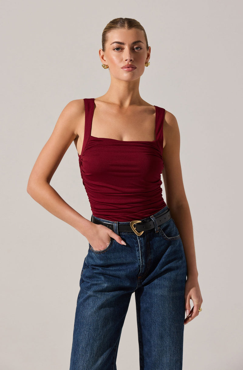 A young woman is wearing a sleeveless, fitted, burgundy top with wide straps and a ruched design across the front. She pairs it with high-waisted, dark-wash jeans and a black belt with a gold heart-shaped buckle.