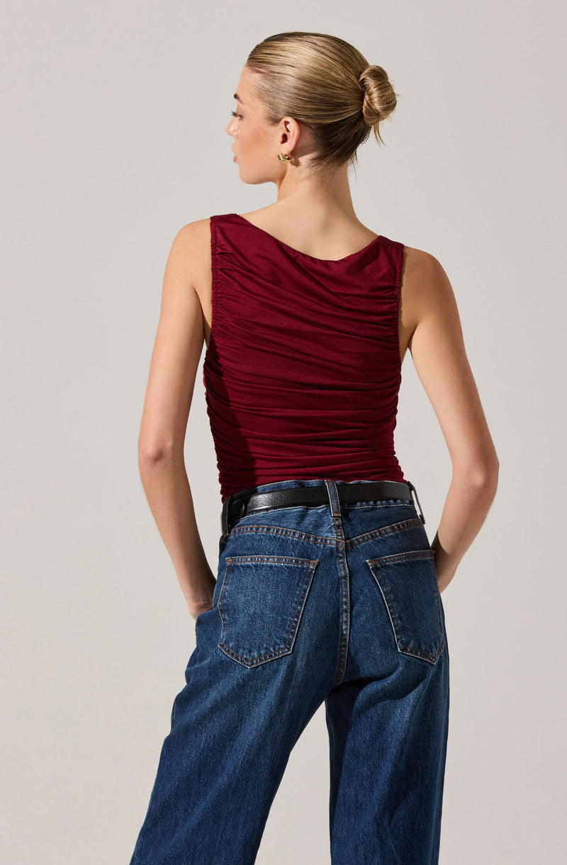 A woman is wearing a sleeveless, burgundy top with gathered, ruched fabric details and a wide neckline. She has paired the top with high-waisted, dark blue jeans and a black belt.