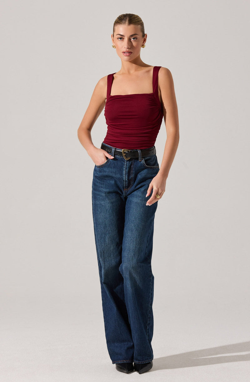 A woman is wearing a fitted, sleeveless, burgundy top with wide shoulder straps and a gathered, ruched design across the chest. She has paired it with high-waisted, dark blue, straight-leg jeans and a black belt with a gold buckle.