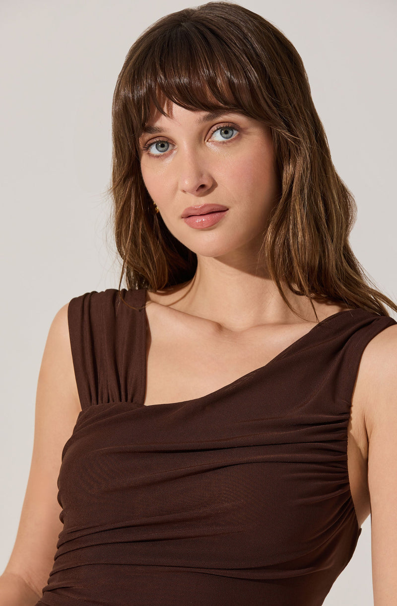 A woman is wearing a sleeveless, dark brown dress with gathered fabric detailing across the bust and shoulders. The dress has wide straps that drape over her shoulders.