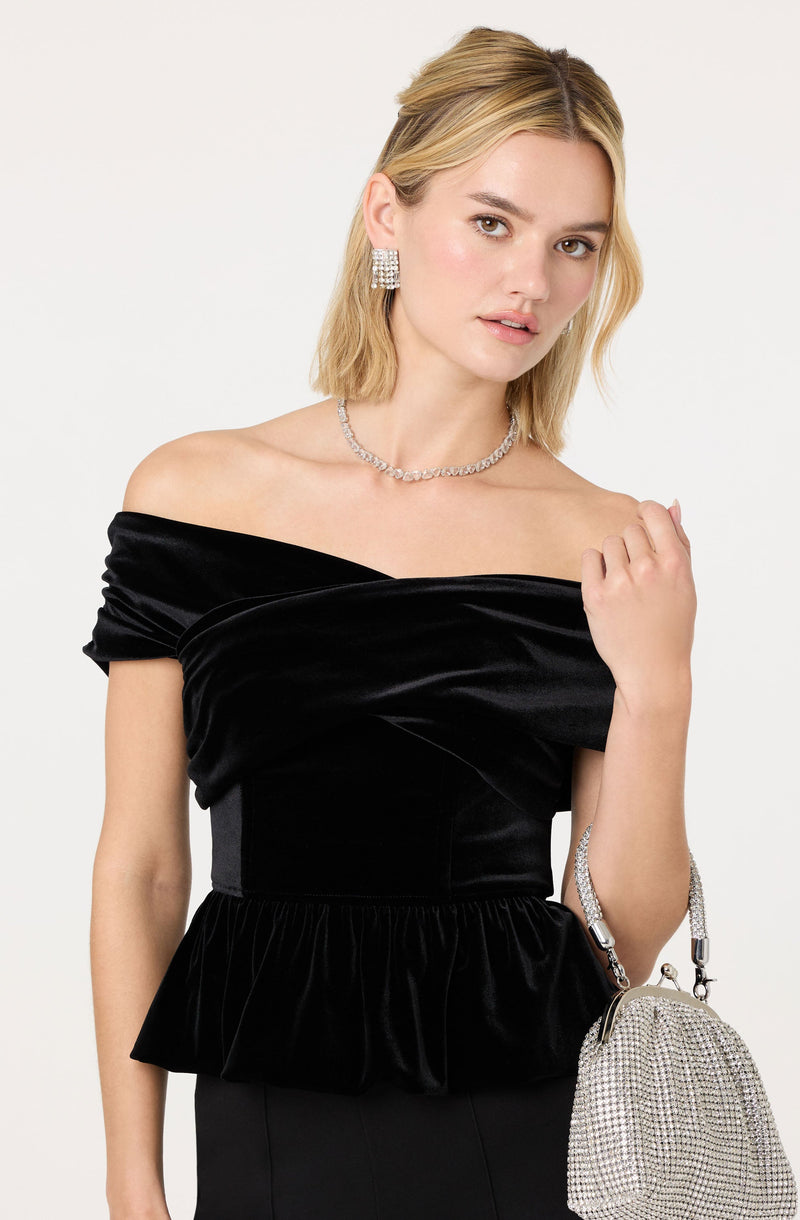 A woman is wearing an elegant off-the-shoulder black velvet top with a ruffled peplum hem. She has accessorized with large, sparkling rectangular earrings, a matching sparkling choker necklace, and a silver rhinestone handbag with a chain strap.