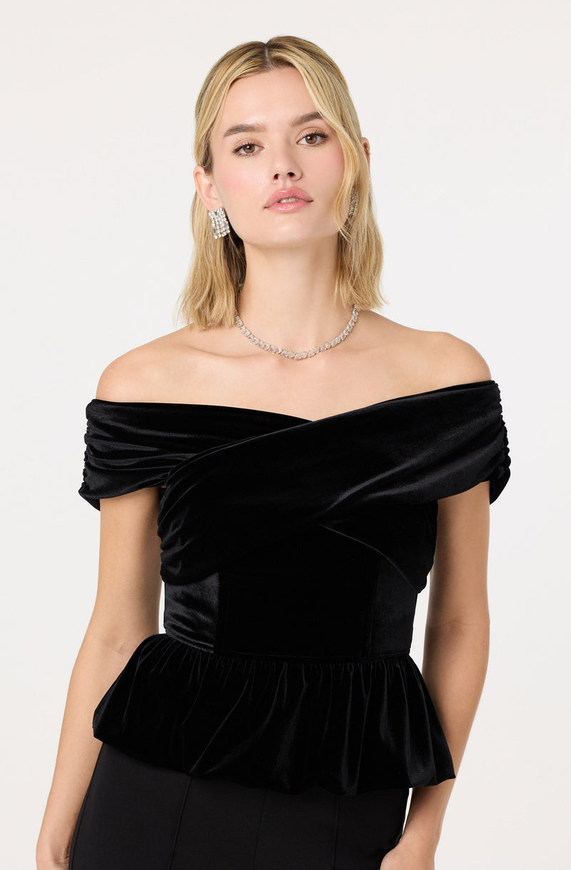 A woman is wearing an off-the-shoulder black dress made of velvet fabric. The dress features a wrapped bodice with a gathered, ruffled hemline at the waist. She accessorizes with large, sparkly earrings and a matching necklace.