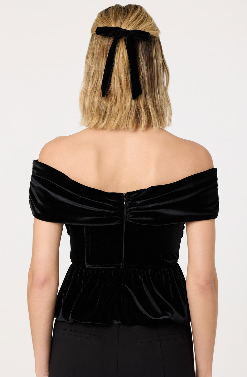 The image shows a woman wearing an off-the-shoulder black velvet top with a ruched design across the shoulders and waist. She has a black ribbon tied in a bow around her head, securing her shoulder-length, wavy blonde hair.