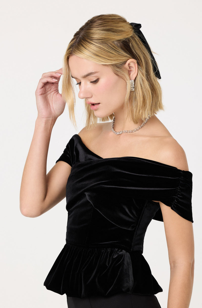 A woman is wearing an elegant off-the-shoulder black velvet top with gathered fabric that creates a ruffled peplum detail at the waist. The top has short, ruched sleeves, and she accessorizes with a pearl necklace, square statement earrings, and a black bow in her short blonde hair.