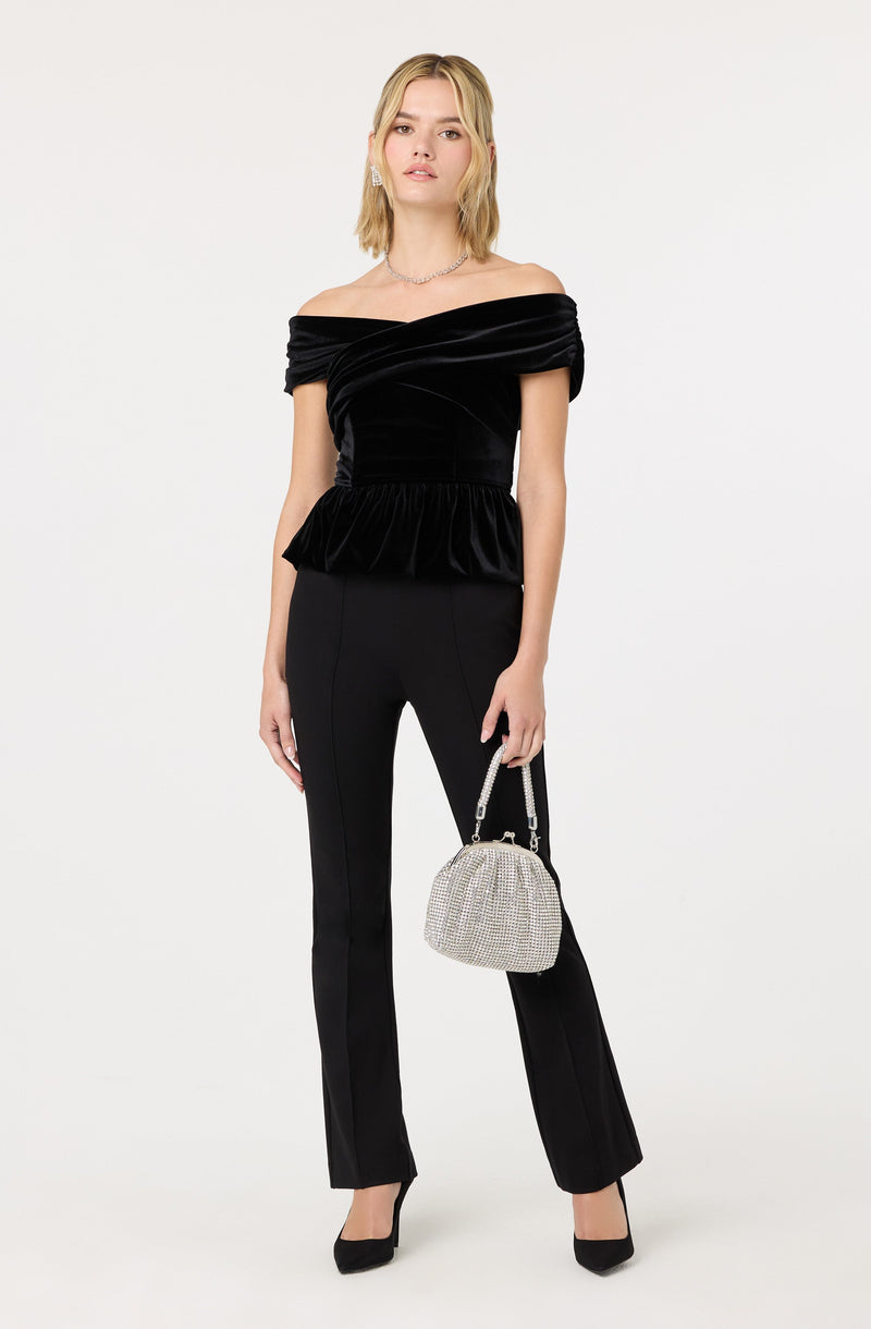 A woman is wearing an off-the-shoulder black velvet top with a ruched bodice and a ruffled peplum waist. She pairs it with black fitted pants that have a slight flare at the hem. She is accessorizing with a small, sparkly silver handbag with a chain strap, black high heels, and a delicate necklace.
