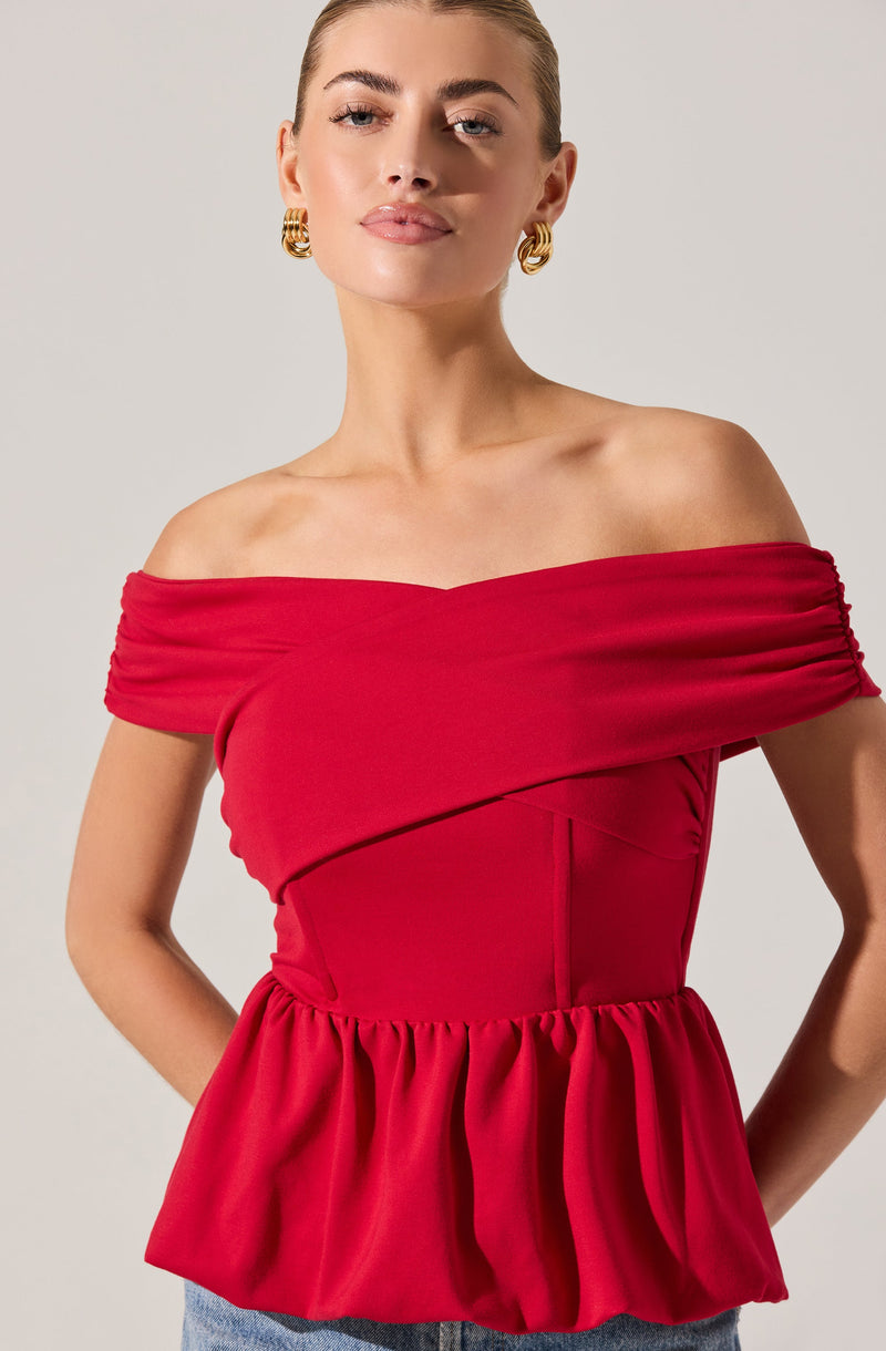 A woman is wearing an off-the-shoulder dress in a bright red color. The dress features a fitted bodice with horizontal ruching along the neckline and a gathered peplum-style waist. She is accessorized with large gold hoop earrings.
