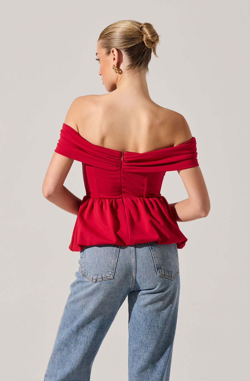 A woman is wearing an off-the-shoulder red top with a fitted bodice and a peplum hem. The top features ruched detailing across the bust and long, draped sleeves that sit off the shoulders. She pairs it with high-waisted, light-wash jeans.