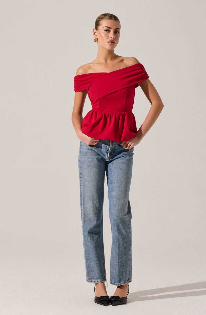 A woman is wearing an off-the-shoulder red top with ruching details across the shoulders and a fitted bodice that flares into a gathered peplum at the waist. She pairs the top with light blue straight-leg jeans and black pointed-toe heels with bows on the front.