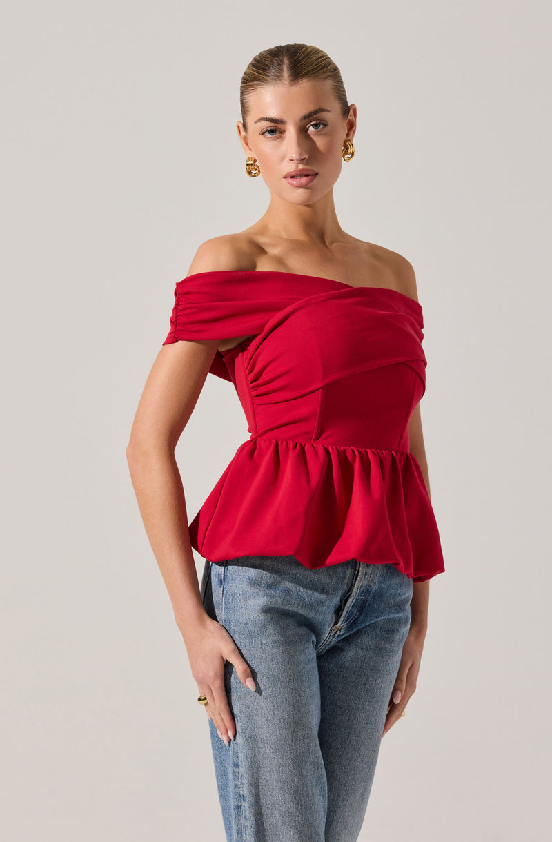 A woman is wearing a bright red, off-the-shoulder top with gathered, ruched detailing at the sleeves and a fitted bodice that extends into a peplum-style hem with a ruffled finish. She pairs the top with light blue jeans. She has gold hoop earrings and is posed against a plain, light-colored background.