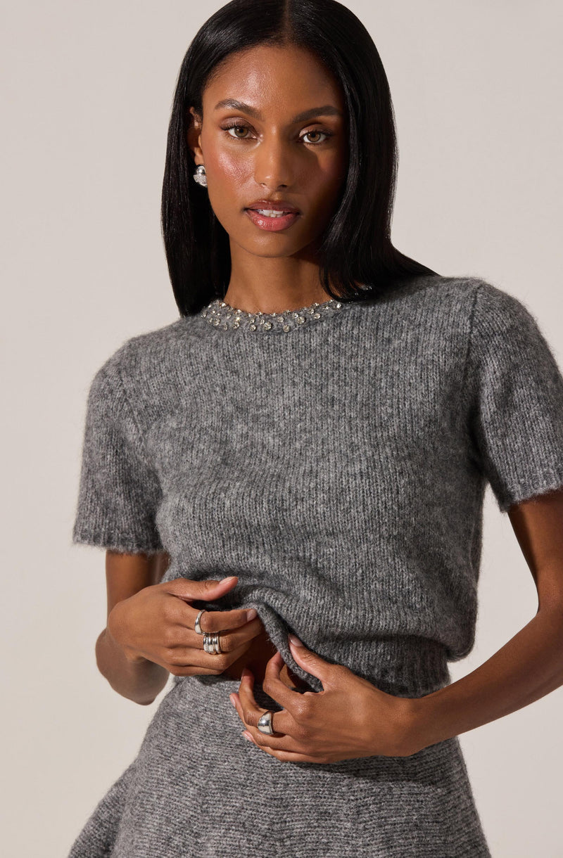 A woman is wearing a short-sleeved, crew neck sweater made of soft, ribbed knit fabric in a heather gray color. The sweater features a decorative embellishment of pearls or beads along the neckline. She pairs it with a matching heather gray skirt. She is accessorized with multiple rings on her fingers and pearl stud earrings.