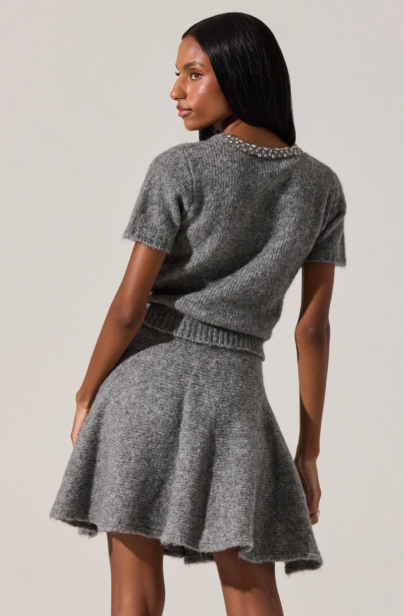 A woman with long, straight black hair is wearing a matching gray knit outfit. The top is a short-sleeved, slightly oversized sweater with a beaded or embellished neckline. The bottom is a high-waisted, flared knit skirt made from the same gray material, creating a cohesive look.