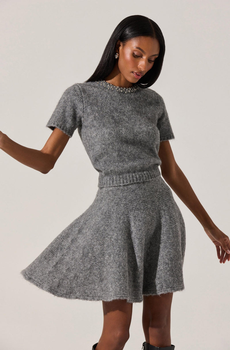 A woman is wearing a gray knit sweater with short sleeves and a round neckline decorated with small embellishments, paired with a matching gray knit skirt that has a high waist and a flared, A-line silhouette.
