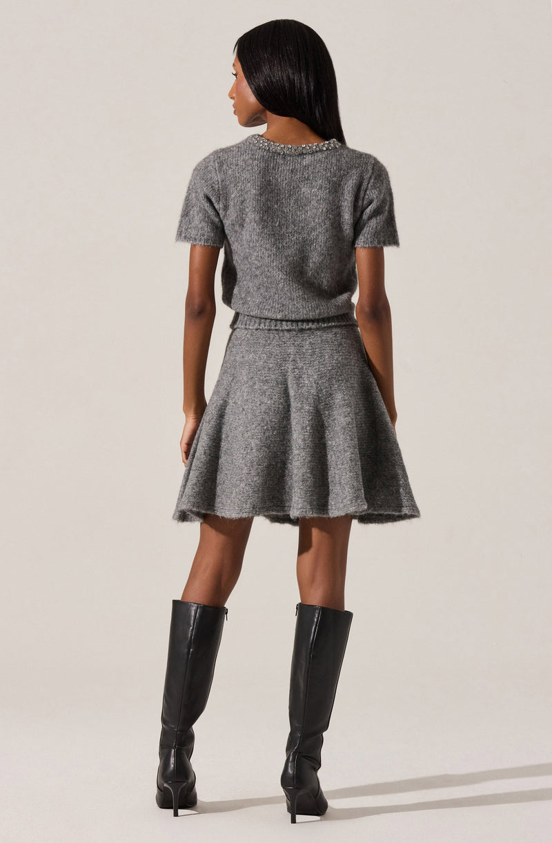 A woman is wearing a matching grey knit short-sleeve sweater and a flared skirt, both made of a soft, textured material. The sweater has a decorative trim around the neckline. She is also wearing tall black leather knee-high boots with high heels.