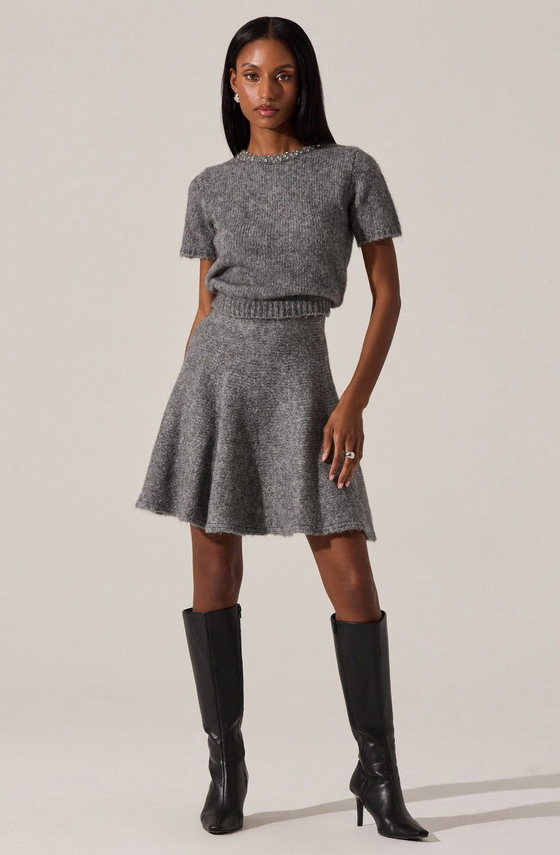 A woman is wearing a matching gray knit outfit consisting of a short-sleeve, slightly loose sweater with a beaded neckline and a high-waisted, flared skirt. She has paired the outfit with knee-high black leather boots that have pointed toes and stiletto heels.