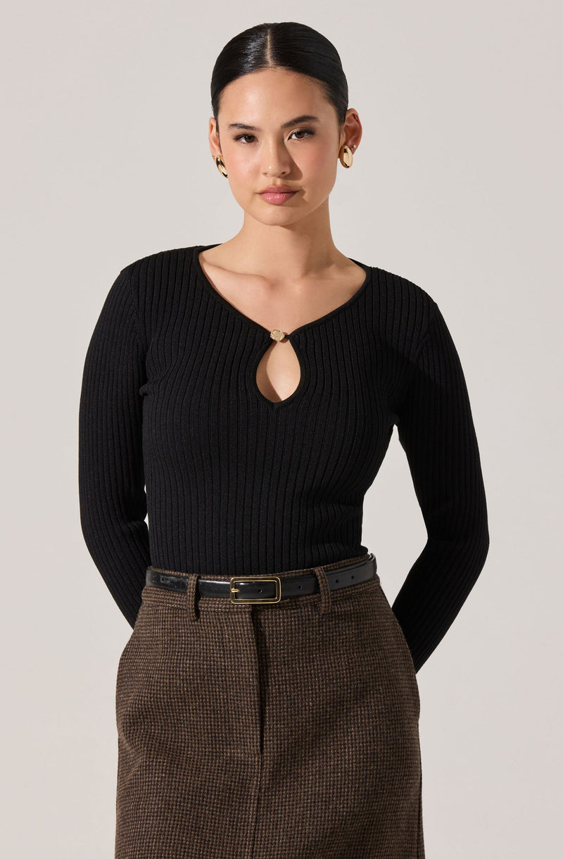 A woman with dark hair styled in a sleek, center-parted manner is wearing a fitted, long-sleeved black ribbed sweater with a small keyhole opening at the neckline, secured by a decorative clasp. She is also wearing high-waisted brown checkered or houndstooth patterned pants with a black belt that has a gold rectangular buckle.