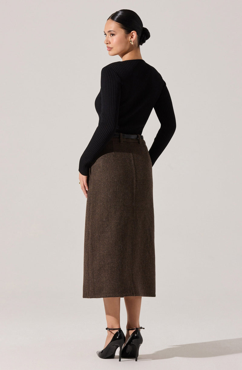A woman is wearing a fitted black long-sleeve ribbed sweater tucked into a high-waisted, ankle-length brown skirt with a subtle checkered pattern. She is also wearing black high-heeled shoes with ankle straps.